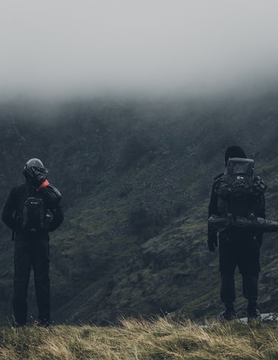 一个穿着黑夹克黑裤子背着黑背包的男人站在绿草覆盖的小山上